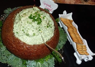 Close-up photo of a rye bread bowl with spinach spread in it. A white tray is next to it filled with crackers.
