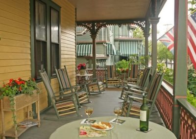 A close-up photo of the Prince Albert Hall porch with rockers lining the porch and the American and British flags flying. The flower box and planter are filled with red flowers and the table is set with white wine and crackers for two.