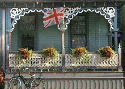 Photo of the Lord Melbourne porch taken from the street. There is a Queen Victoria bicycle parked in front of the porch. The flower boxes are overflowing with greenery and the British flag is swaying in the wind.