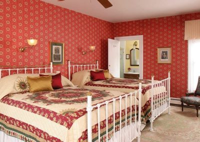 Prince Albert guestroom with rich red wallpaper and two queen beds with white iron bed frames. Beds have traditional red, gold and green quilts