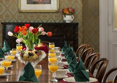 The Queen Victoria Dining Room Table set for breakfast with orange juice at each place setting. A bowl of fresh cut fruit in a glass bowl is on the table. A vase of multi-color tulips is the centerpiece.